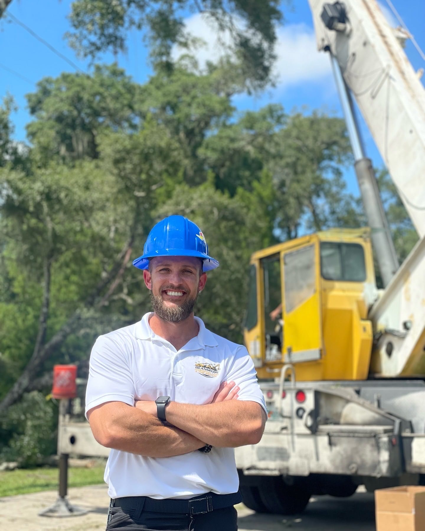 DeAngelo's Land Services owner in blue hard hat and polo standing in front of a yellow crane truck.