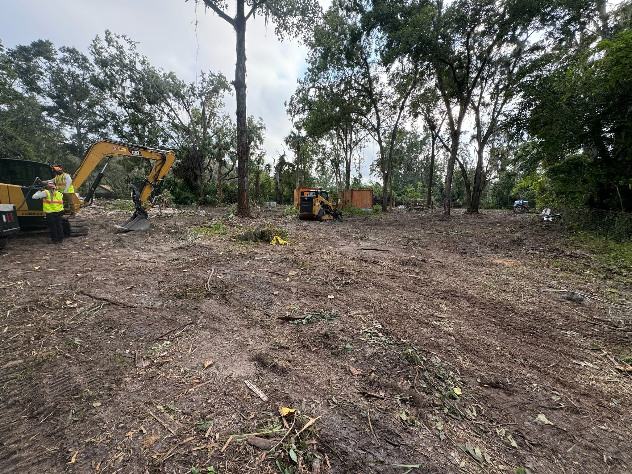 Two CAT excavators and a hi-vis groundsman clearing a Florida lot while preserving the perimeter oaks.